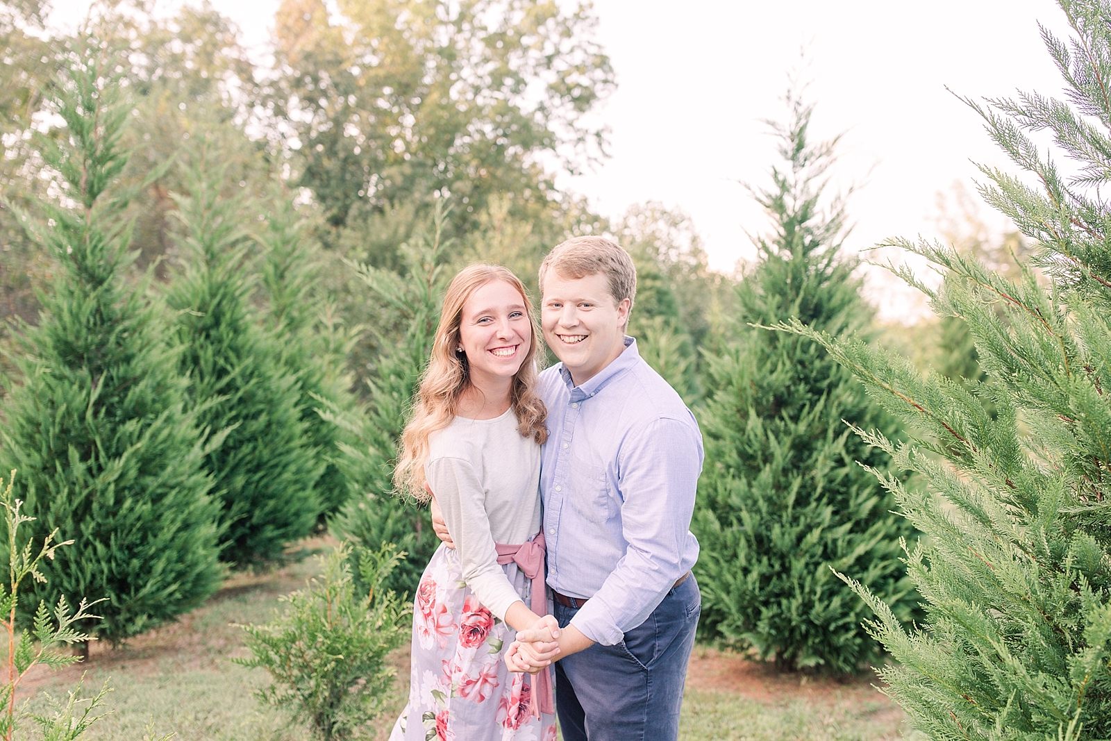 Charlotte Engagement Couple holding hands and smiling at camera surrounded by Christmas trees Photo