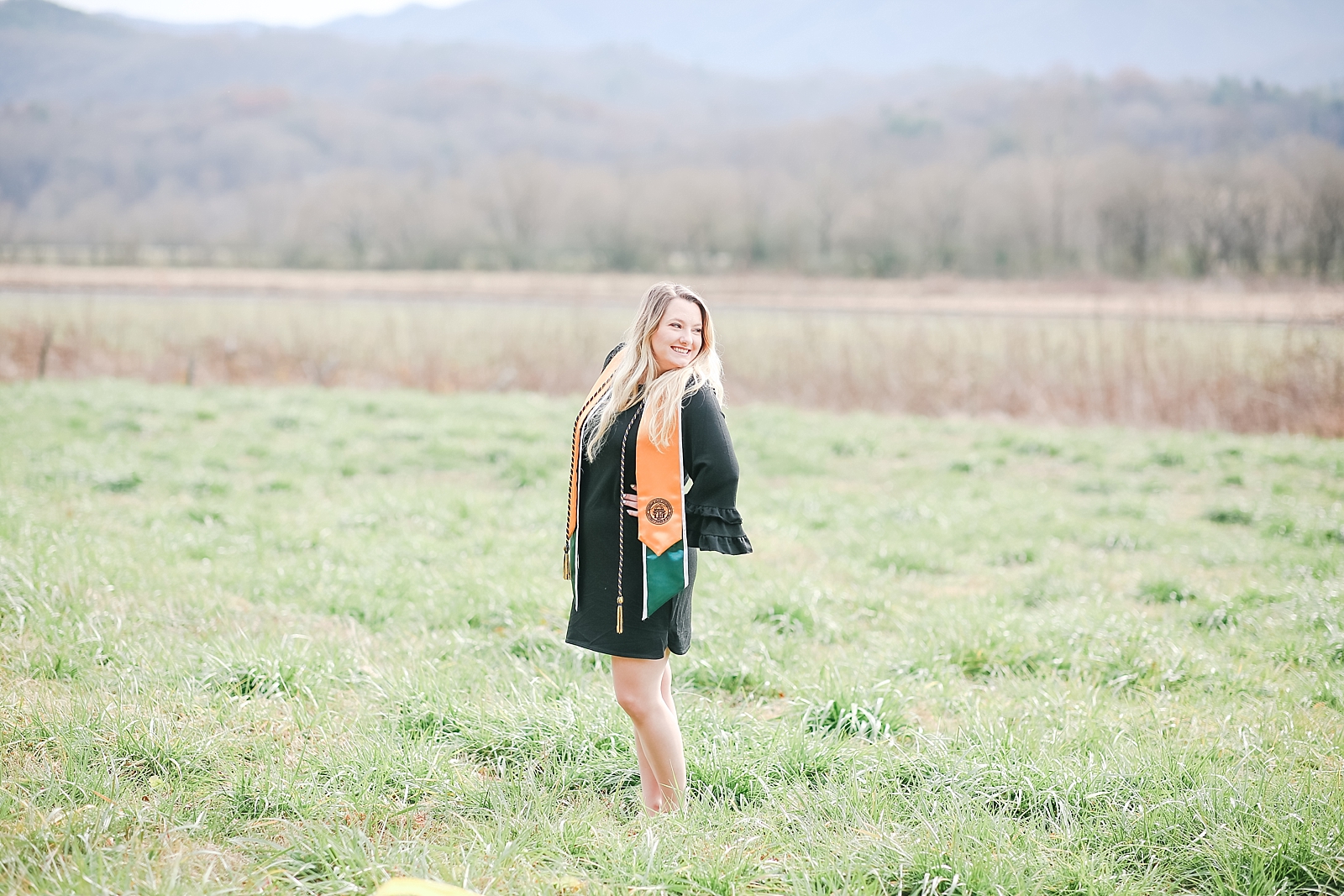 Kennesaw State University Senior Session Rachel in graduation gown standing in field with mountains in the background smiling over her shoulder photo