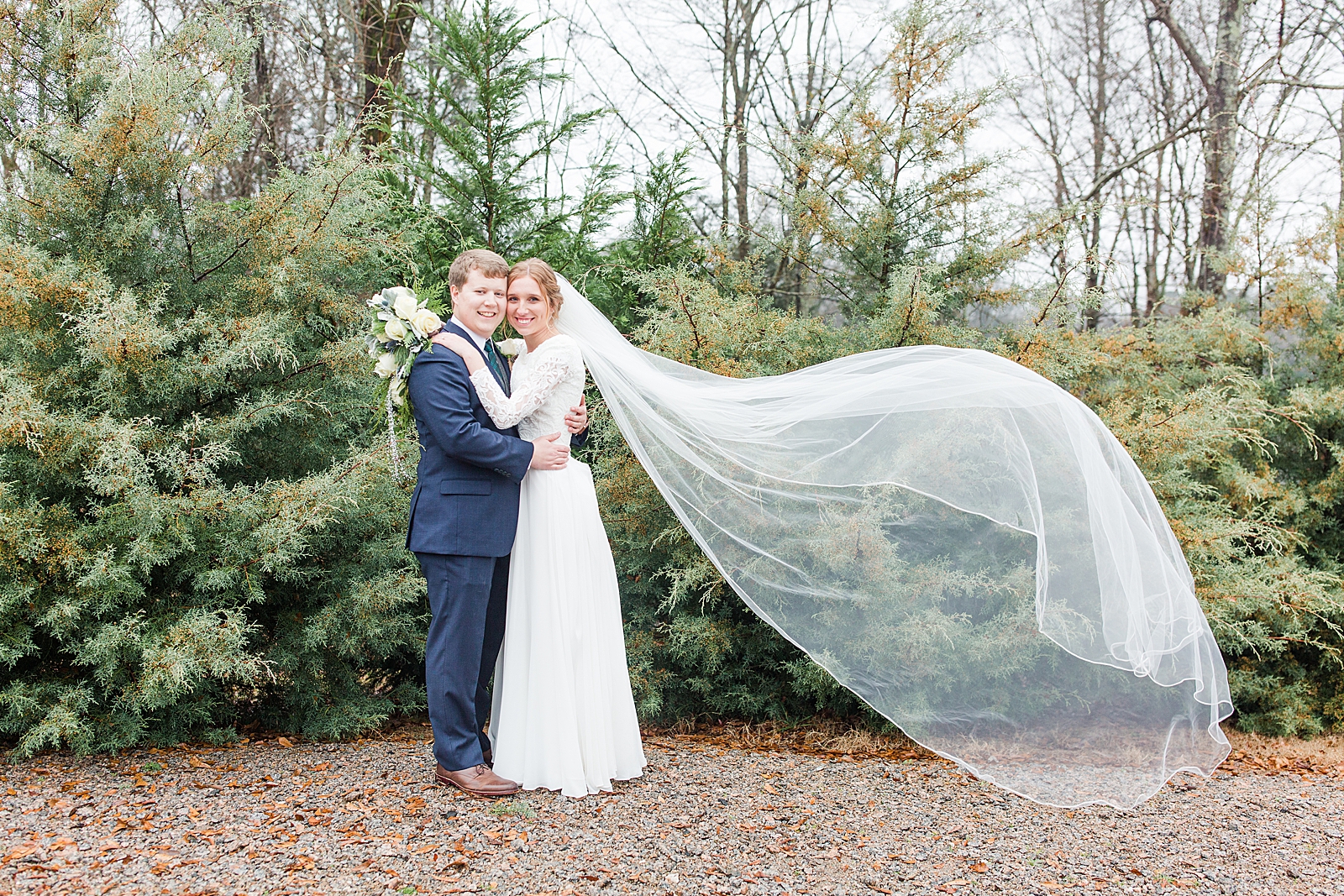 Charlotte Wedding Bride and Groom Smiling at Camera in front of Evergreens with veil blowing Photo