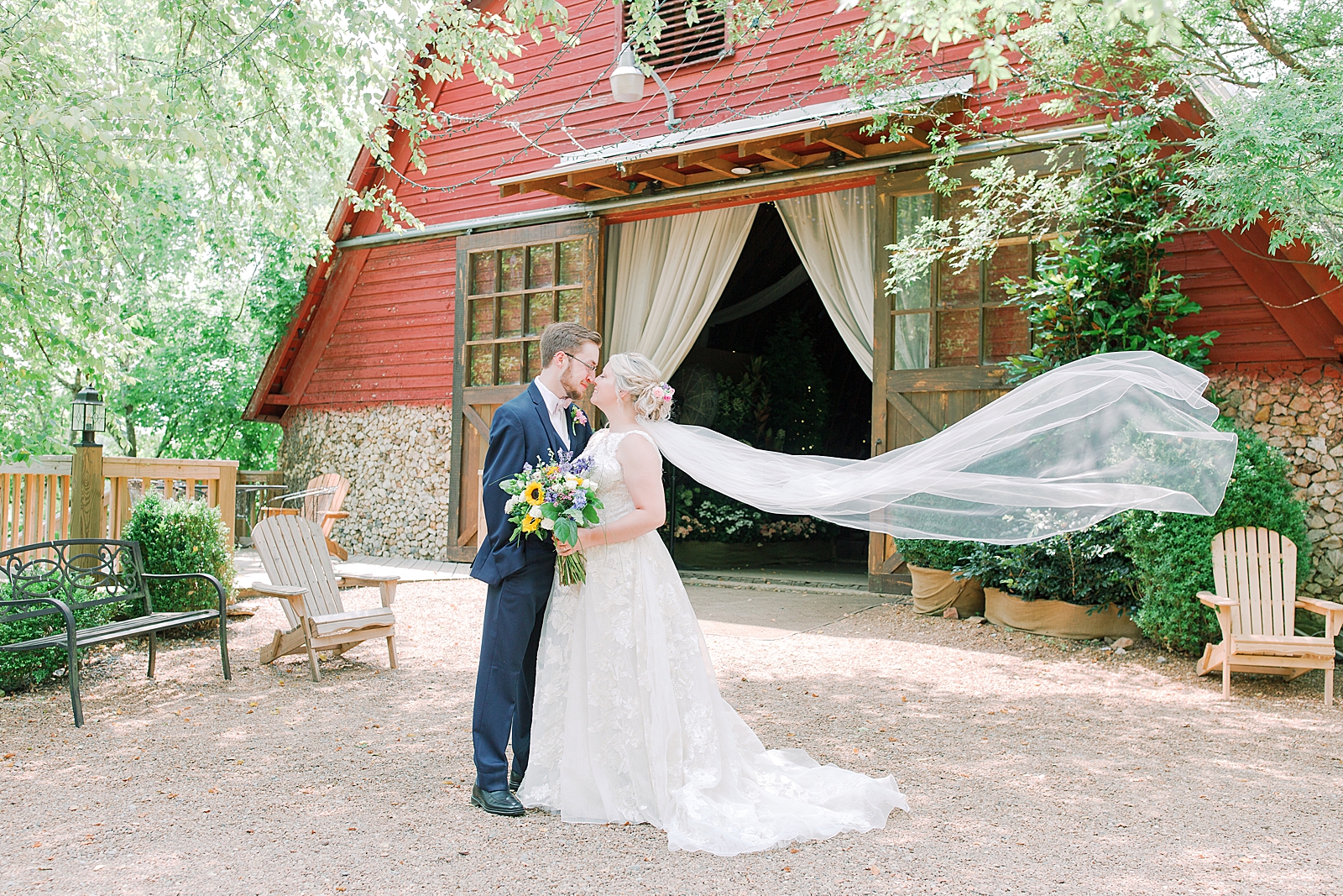 Black Fox Farms Garden Wedding Bride and Groom in Front of Red Barn with Veil Flying Photo