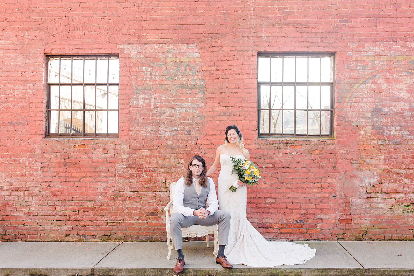 Hackney Warehouse Wedding Bride Standing beside Groom Sitting on Chair in front of brick wall Photo