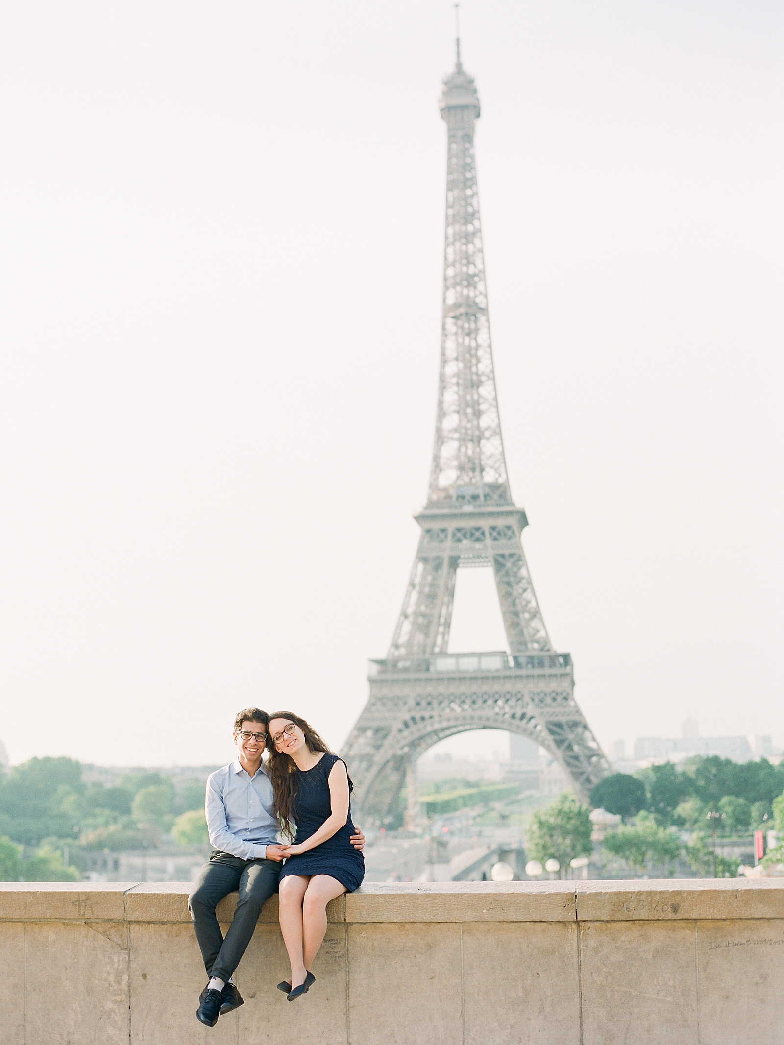Eiffel Tower Engagement Session Couple smiling at camera sitting on wall Photo