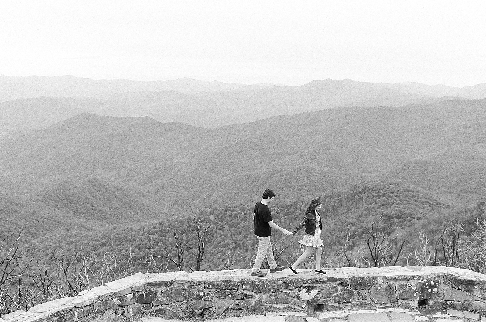 Mountaintop Engagement Black and White Couple walking along wall with mountains in background Photo 