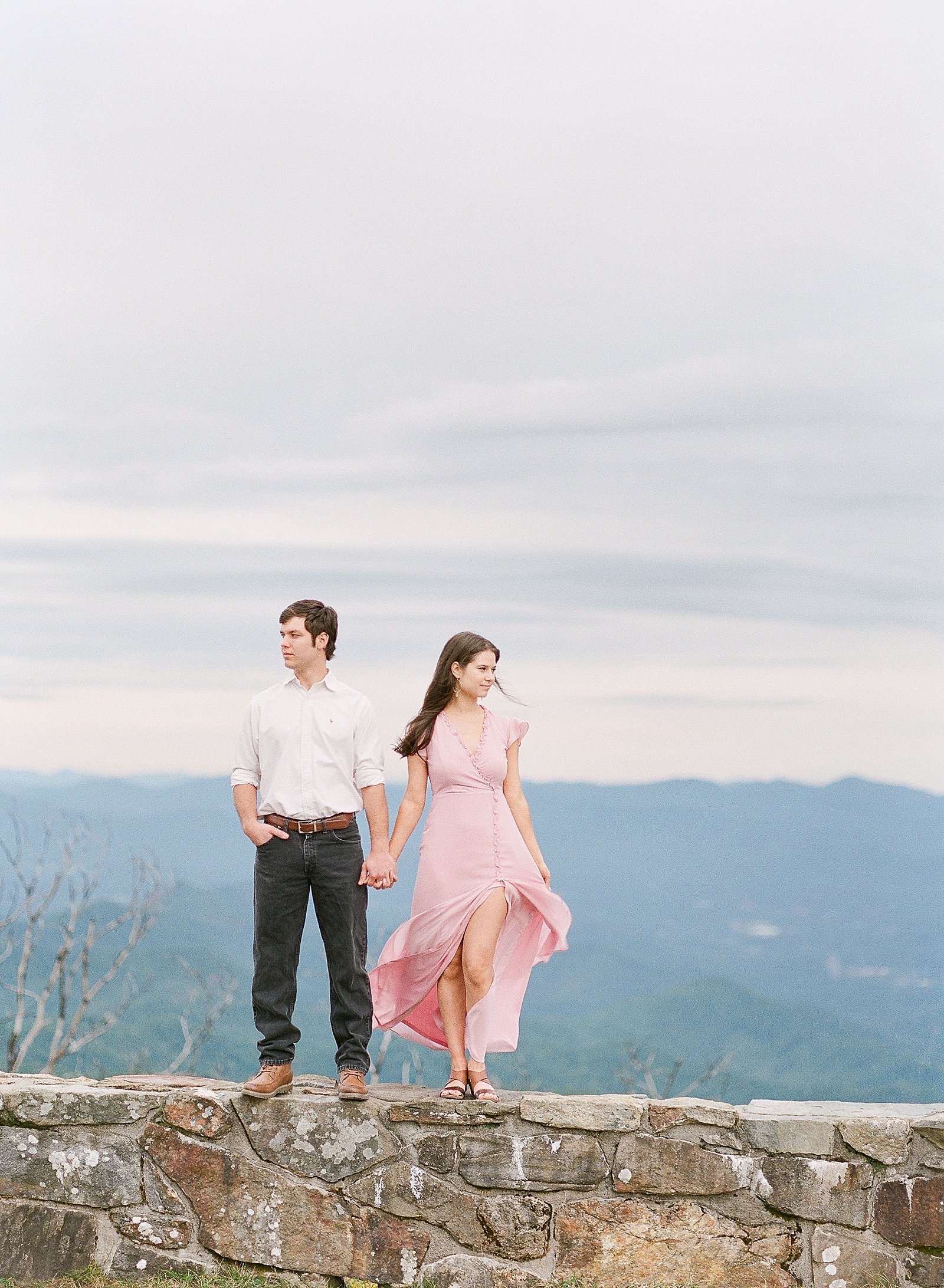 Mountaintop Engagement Couple holding hands looking off standing on rock wall Photo 