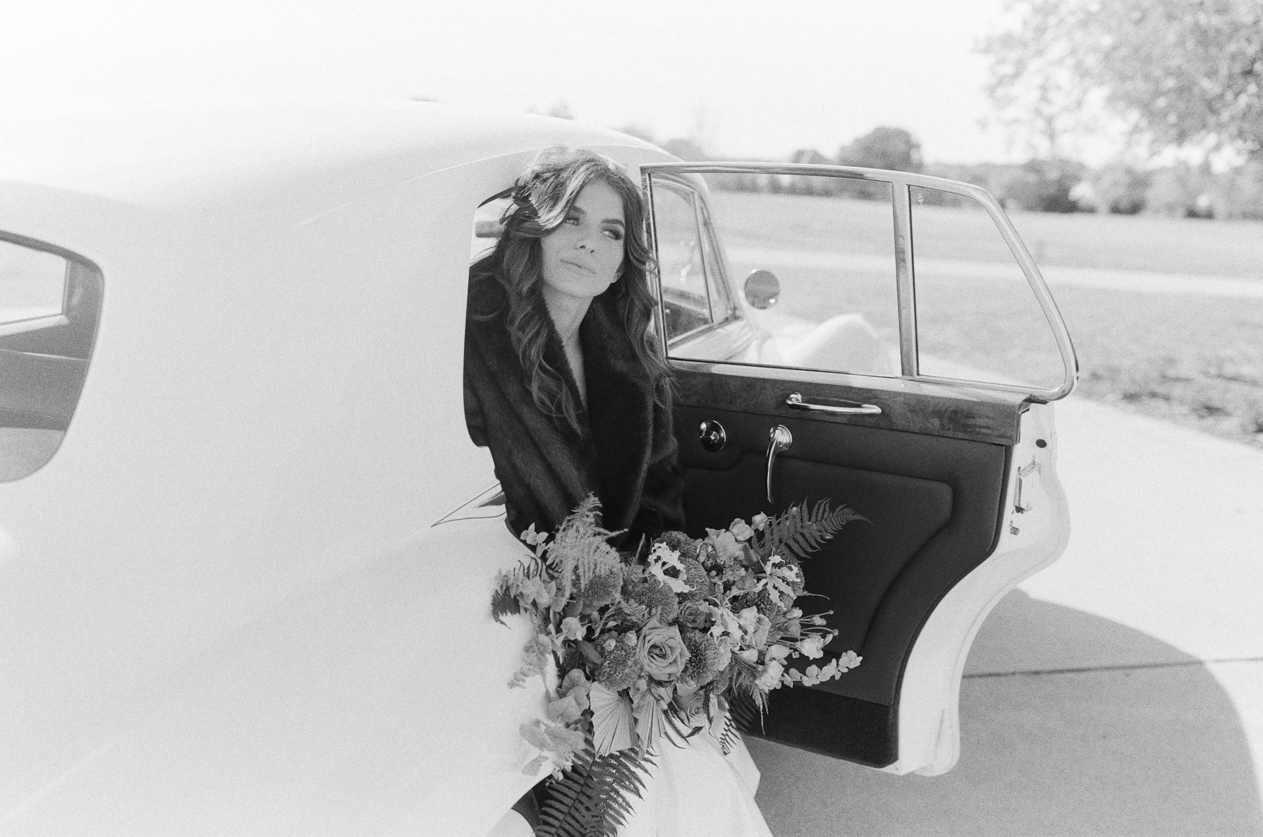 Black and White of Bride in back of Vintage Car Holding Bouquet Photo