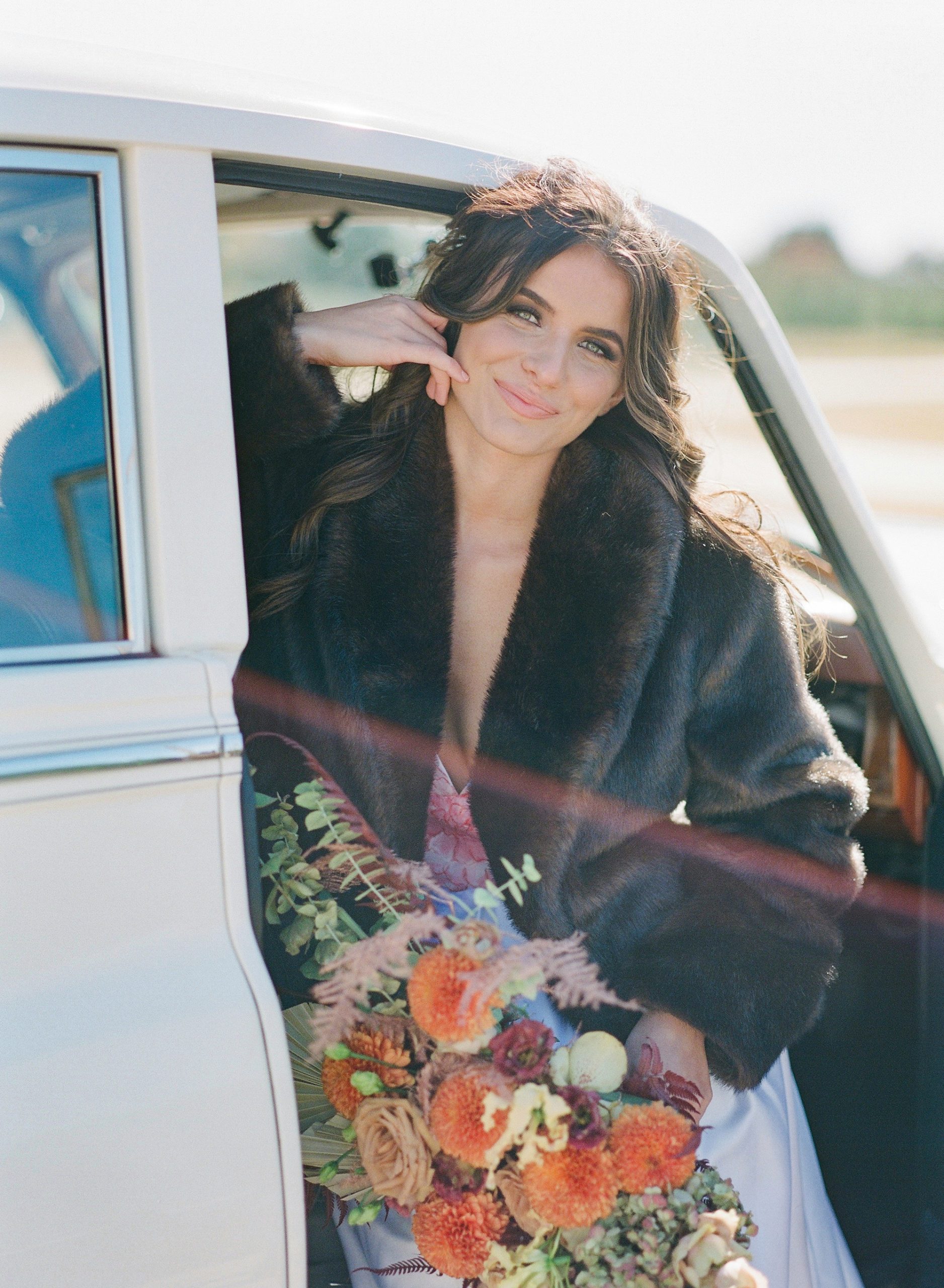 Bride Sitting in Car Smiling at Camera with Hand on her face holding Bouquet Photo 