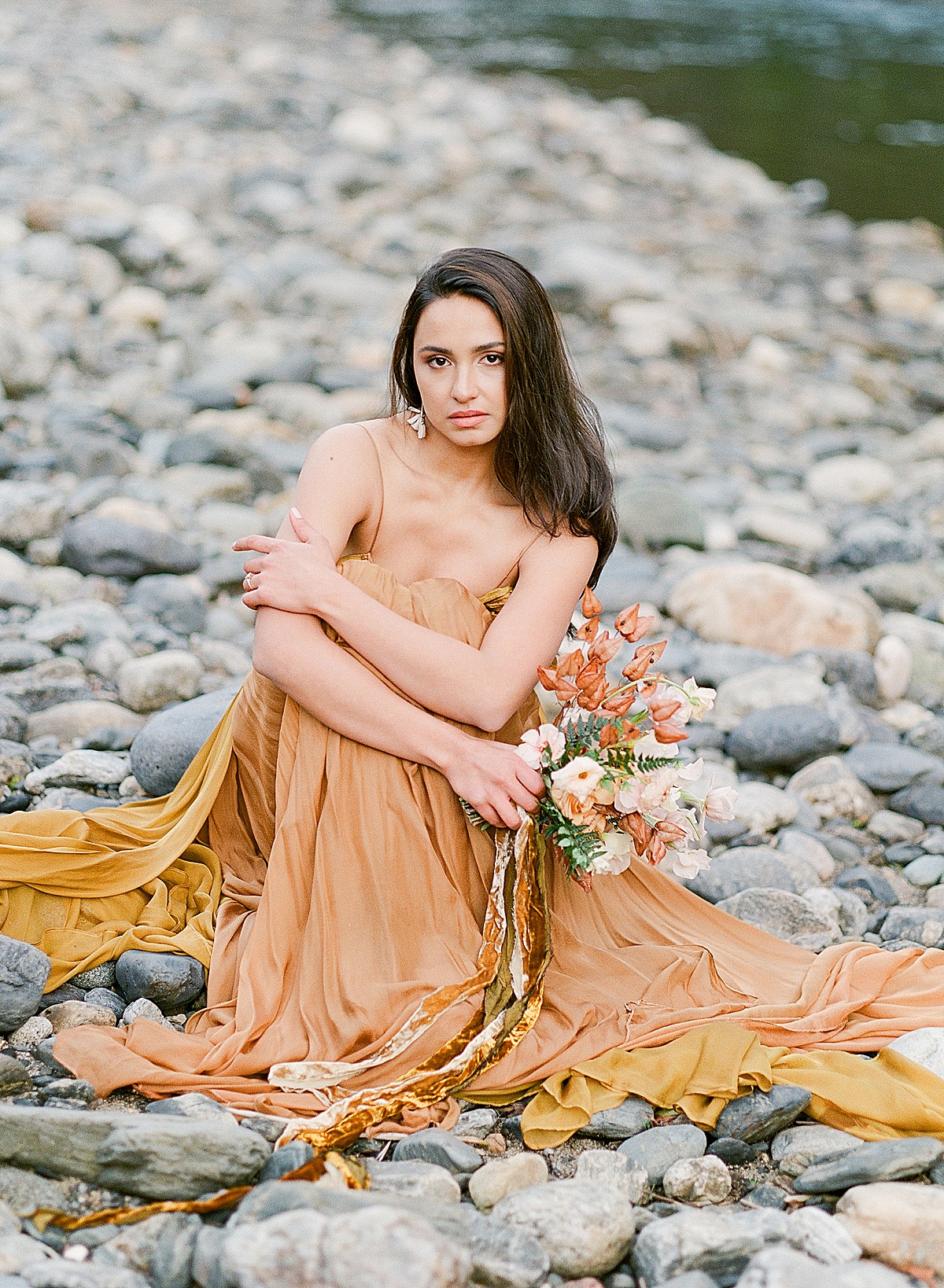 Leanne Marshall Golden Gown Model Sitting On Rocks Holding Bouquet Beside River Photo