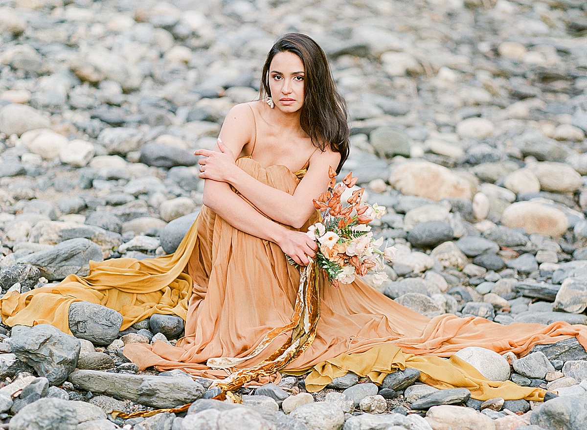Leanne Marshall Golden Gown Model Sitting On Rocks Holding Bouquet Photo