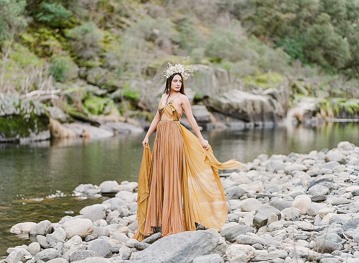Leanne Marshall Golden Gown Model Looking Off Holding Dress Next To River Photo