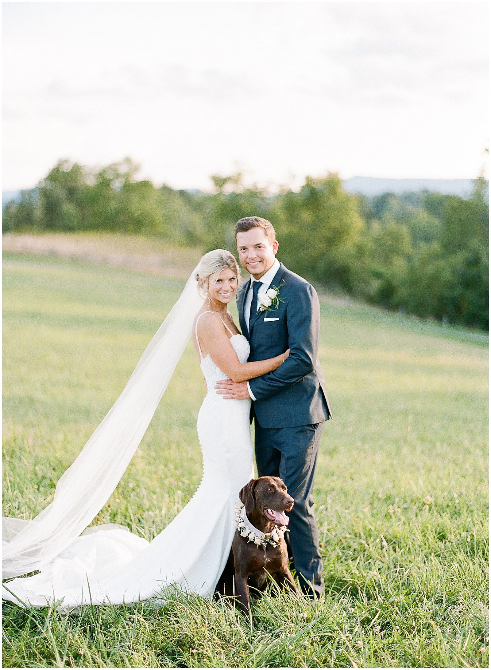 Bride and Groom Smiling at Camera in Field with Their Dog Photo