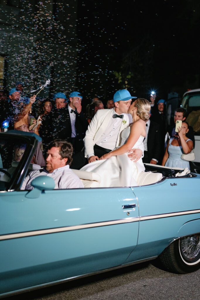Bride and Groom kissing as they leave their wedding reception in a vintage blue convertible.
